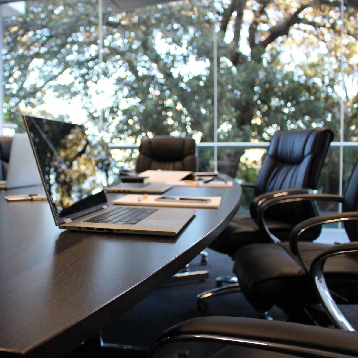 Open laptop computer on large Conference table in front of tall windows with trees outside